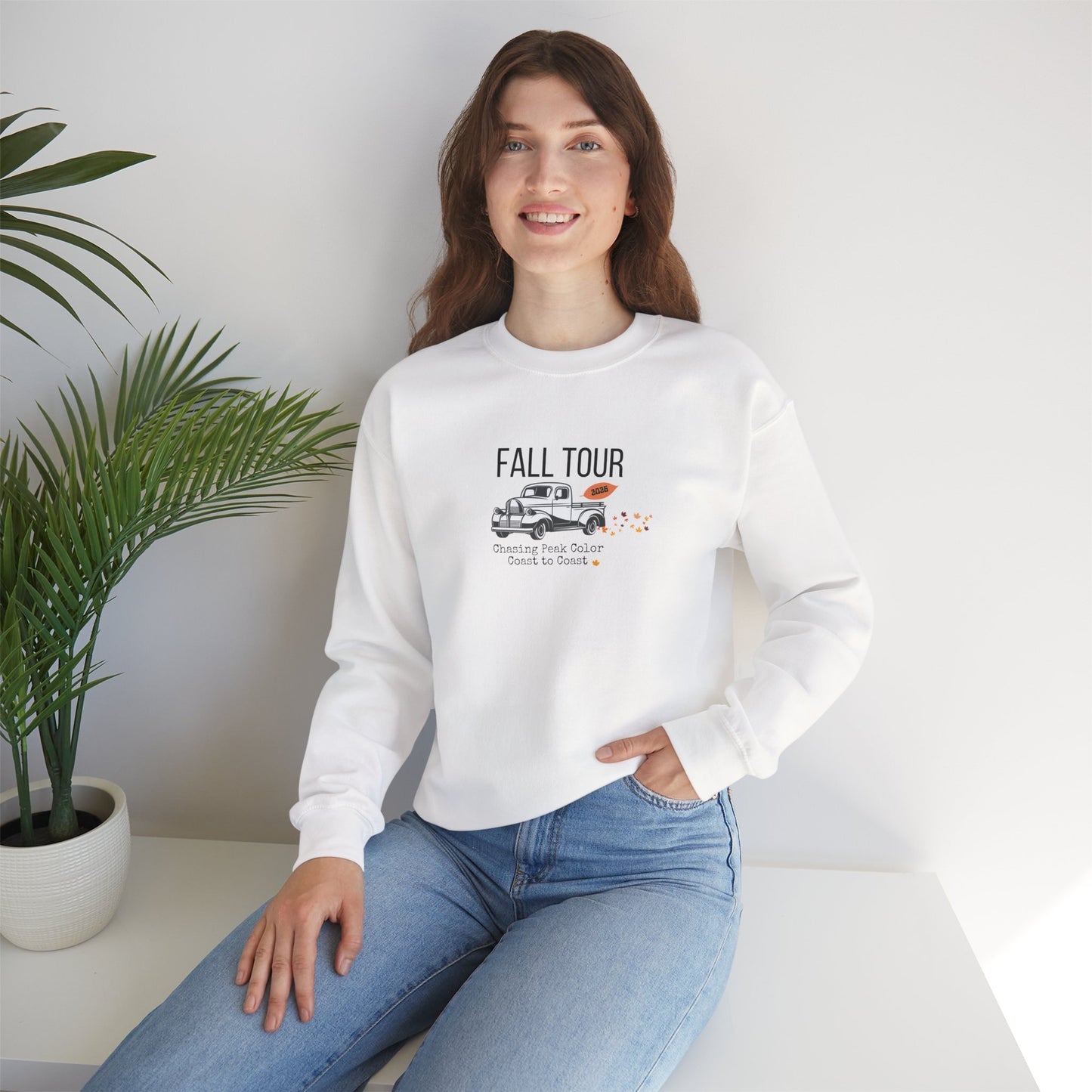 Woman wearing a white sweatshirt with Fall leaf tour printed on it, sitting in a room with a plant.