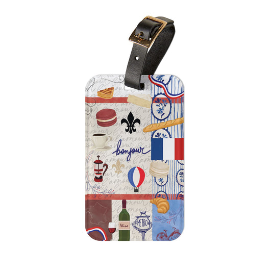A rectangular luggage tag with a black leather strap and gold-tone buckle, featuring a collage-style French-themed print with macarons, croissants, a French flag, a fleur-de-lis, and the word "bonjour" on a white background.