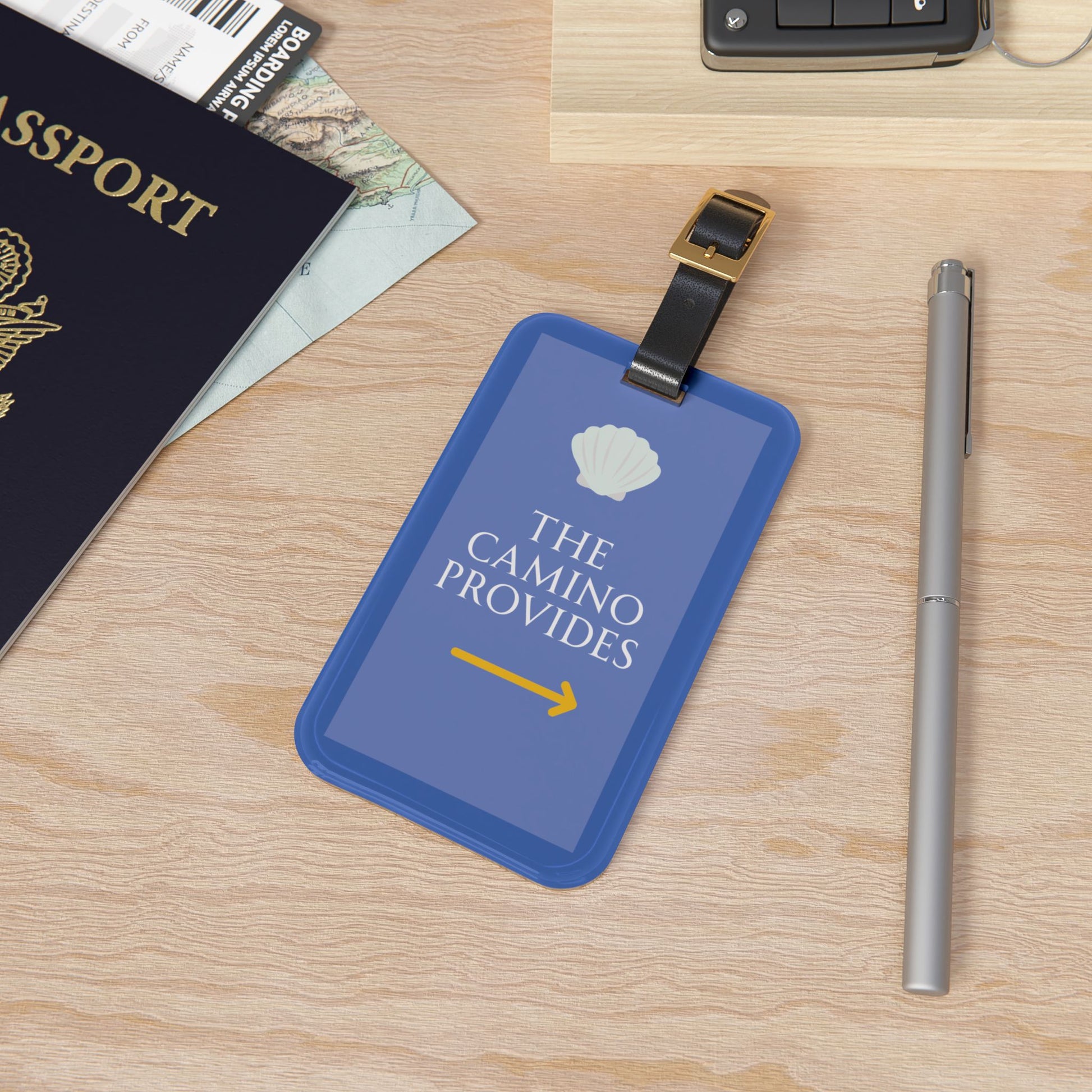Blue luggage tag with 'The Camino Provides' text on a wooden surface with passport and pen.