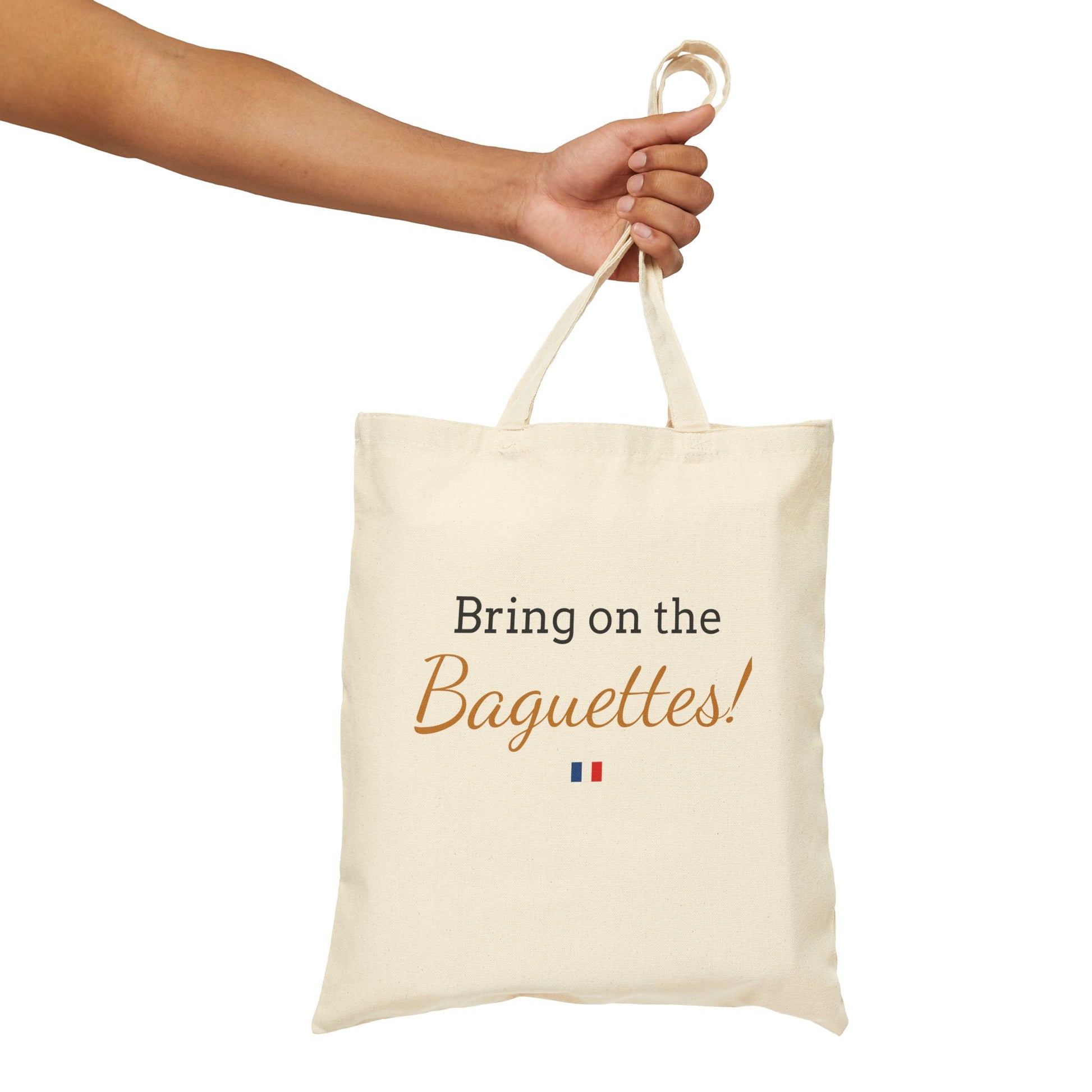 Beige tote bag with 'Bring on the Baguettes!' text held by a hand on a white background