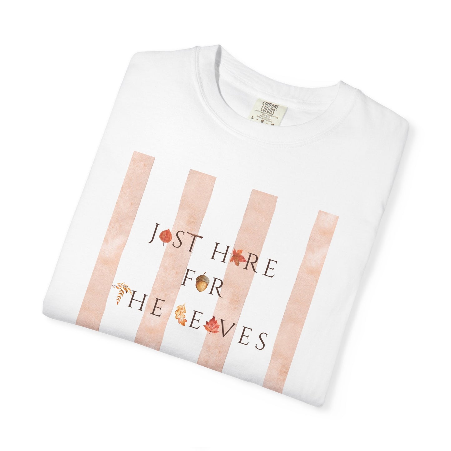 A folded white crewneck t-shirt with a graphic print featuring vertical peach watercolor stripes and the text 'JUST HERE FOR THE EYES' decorated with small autumn leaf and acorn illustrations.