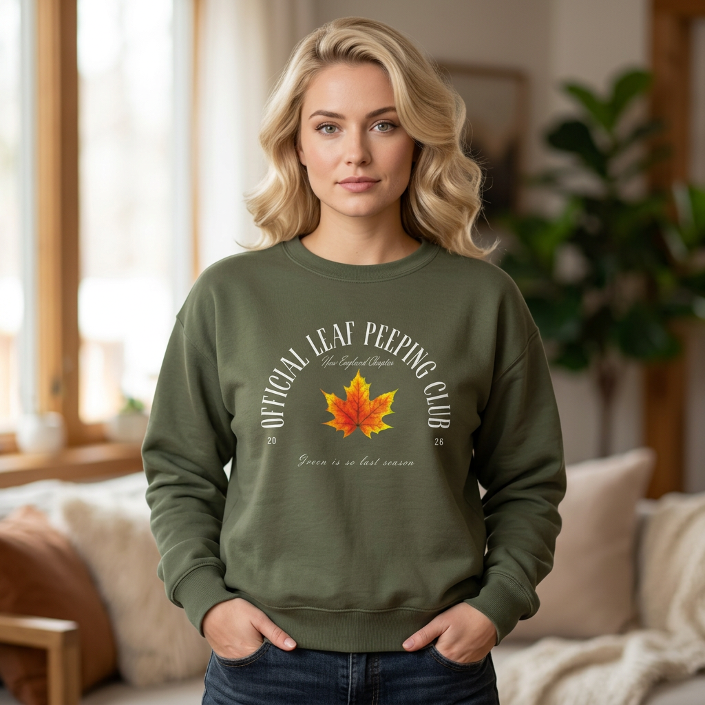 A blonde woman wearing an olive green crewneck sweatshirt with a graphic that reads 'OFFICIAL LEAF PEERING CLUB' around a red and orange maple leaf, standing in a cozy living room setting.