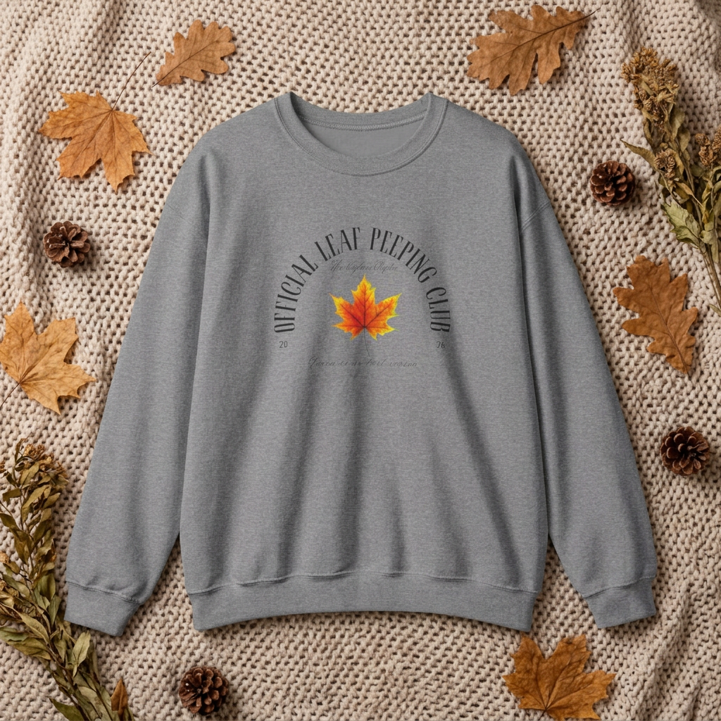 A heather gray crewneck sweatshirt with a graphic print of a red and yellow maple leaf surrounded by autumn leaves and pinecones, laid flat on a beige knit blanket.