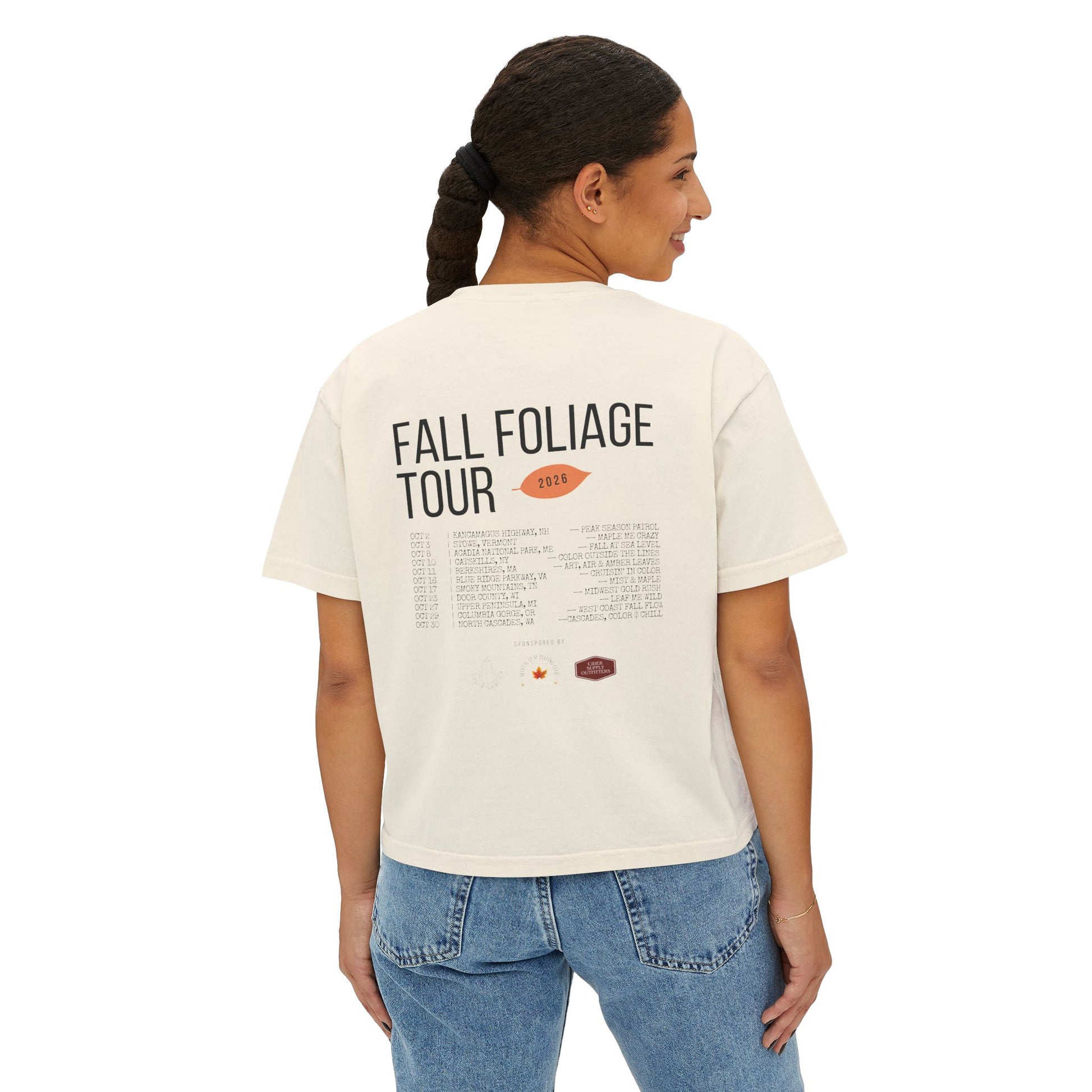 A woman viewed from the back wearing a cream-colored graphic t-shirt with 'FALL FOLIAGE TOUR 2023' printed on the back, paired with blue jeans, against a plain white background.