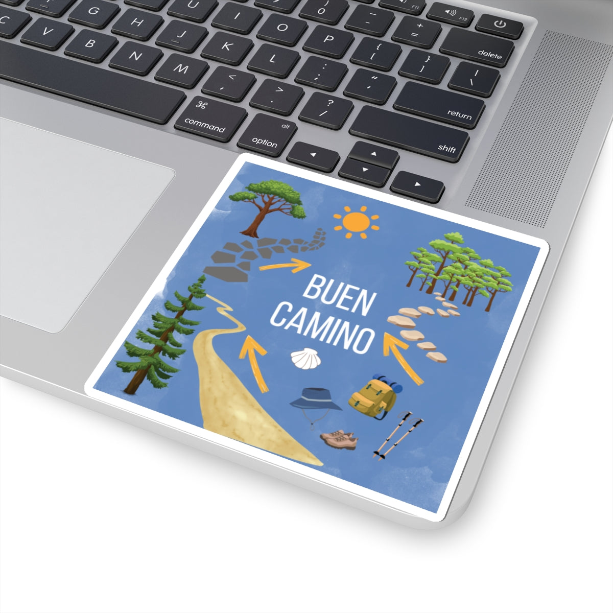 Laptop with a sticker featuring 'Buen Camino' design on a blue background with trees, sun, and arrows.