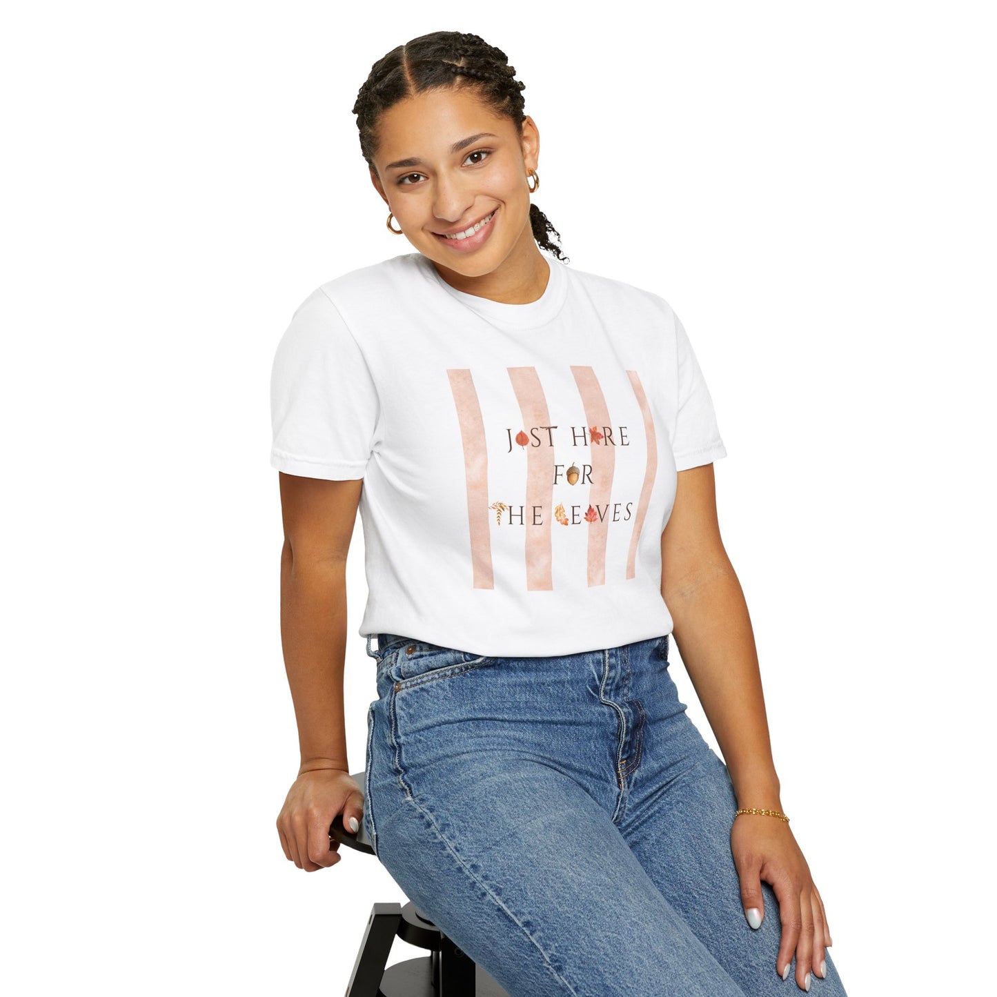 A woman wearing a white graphic t-shirt with vertical pink stripes and the text 'I'M NOT HERE FOR THE LEAVES', paired with blue jeans, seated against a plain white background.