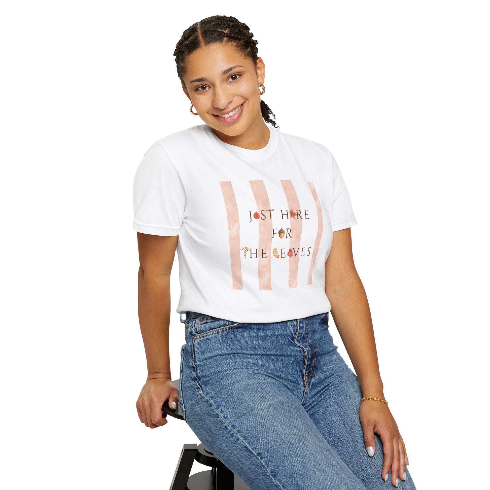 A woman wearing a white graphic t-shirt with vertical pink stripes and the text 'I'M NOT HERE FOR THE LEAVES', paired with blue jeans, seated against a plain white background.