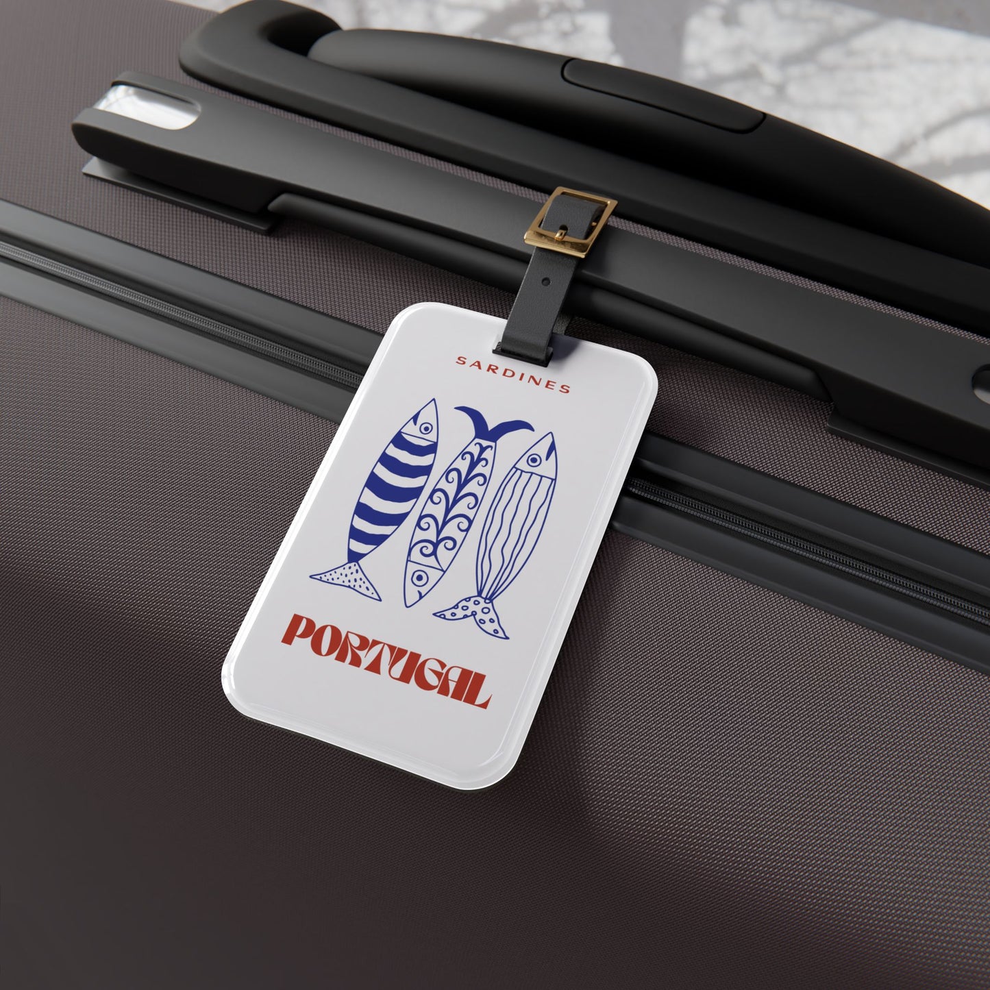 A rectangular white luggage tag with a colorful Mexico-themed illustration and the word Portugal  attached by a black strap with a gold buckle to a dark brown hard-shell suitcase.