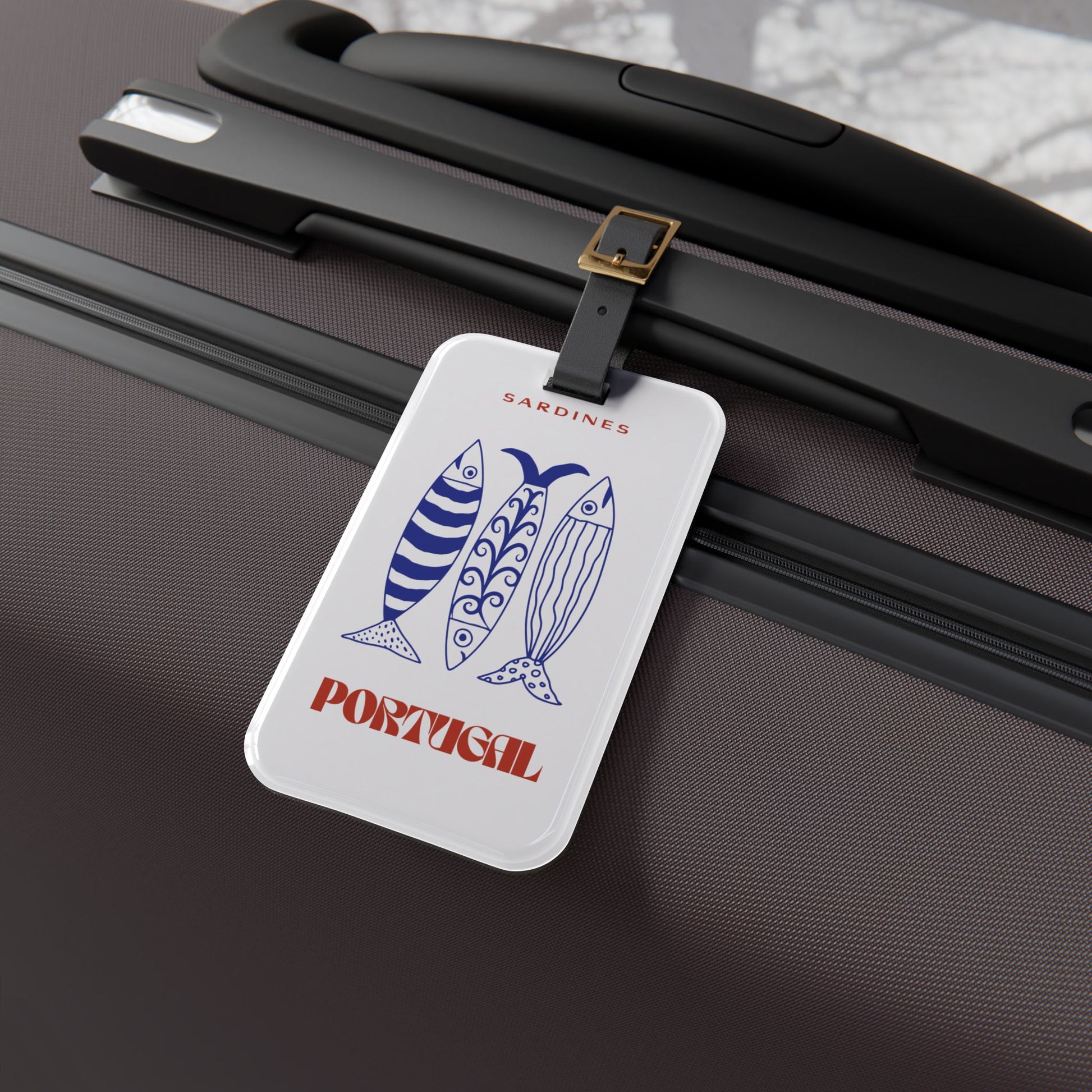 A rectangular white luggage tag with a colorful Mexico-themed illustration and the word Portugal  attached by a black strap with a gold buckle to a dark brown hard-shell suitcase.