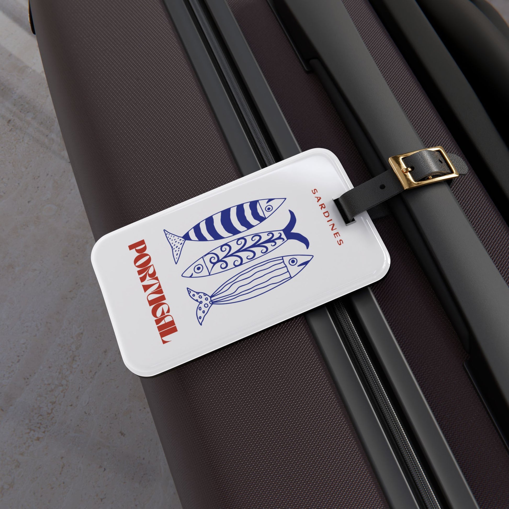 A white luggage tag with a blue striped fish illustration and red text reading 'PORTUGAL and 'SARDINES', attached to a dark brown suitcase.
