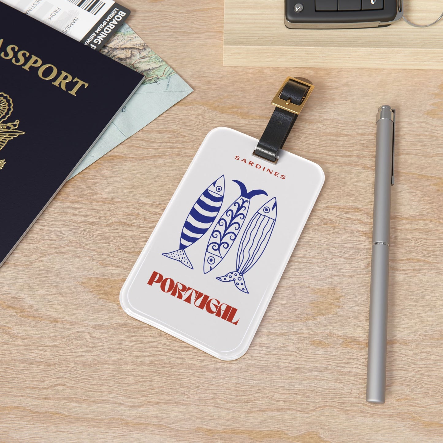 A white luggage tag with a black strap and gold buckle, printed with two blue sardines and the words 'SARDINES' and 'PORTUGAL', placed on a light wood desk next to a passport, pen, and phone.