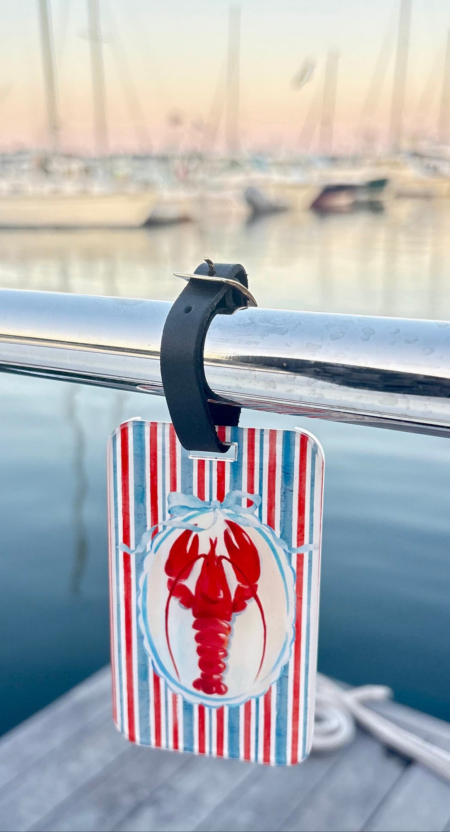A rectangular luggage tag with red, white, and blue vertical stripes and a red lobster illustration in the center, hanging from a black clip on a silver metal railing at a dock with a marina in the background.