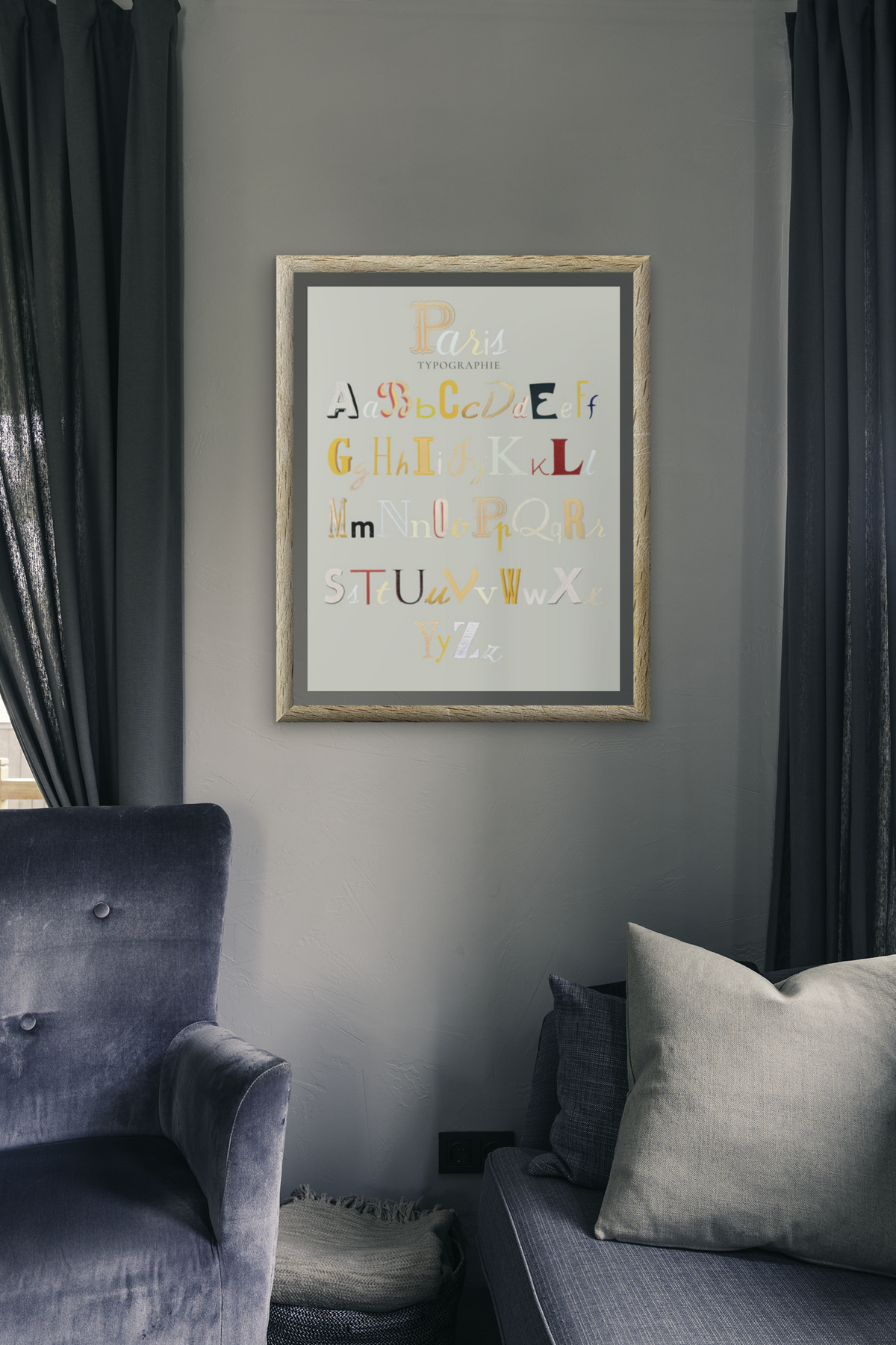 A framed alphabet art print featuring colorful decorative letters on a light gray background, hanging on a gray wall in a styled living room with blue-gray armchairs and dark curtains.