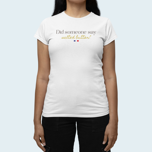A woman wearing a white short-sleeve crewneck t-shirt with the text 'Did someone say salted butter?' printed on the chest in black and gold, with a small French flag graphic underneath. She is also wearing black pants, set against a solid light blue background.