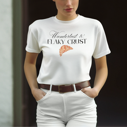 A woman wearing a white crewneck t-shirt with a graphic print that reads 'Moulin & FLAKY CRUST' above a croissant illustration, paired with white pants and a brown belt.
