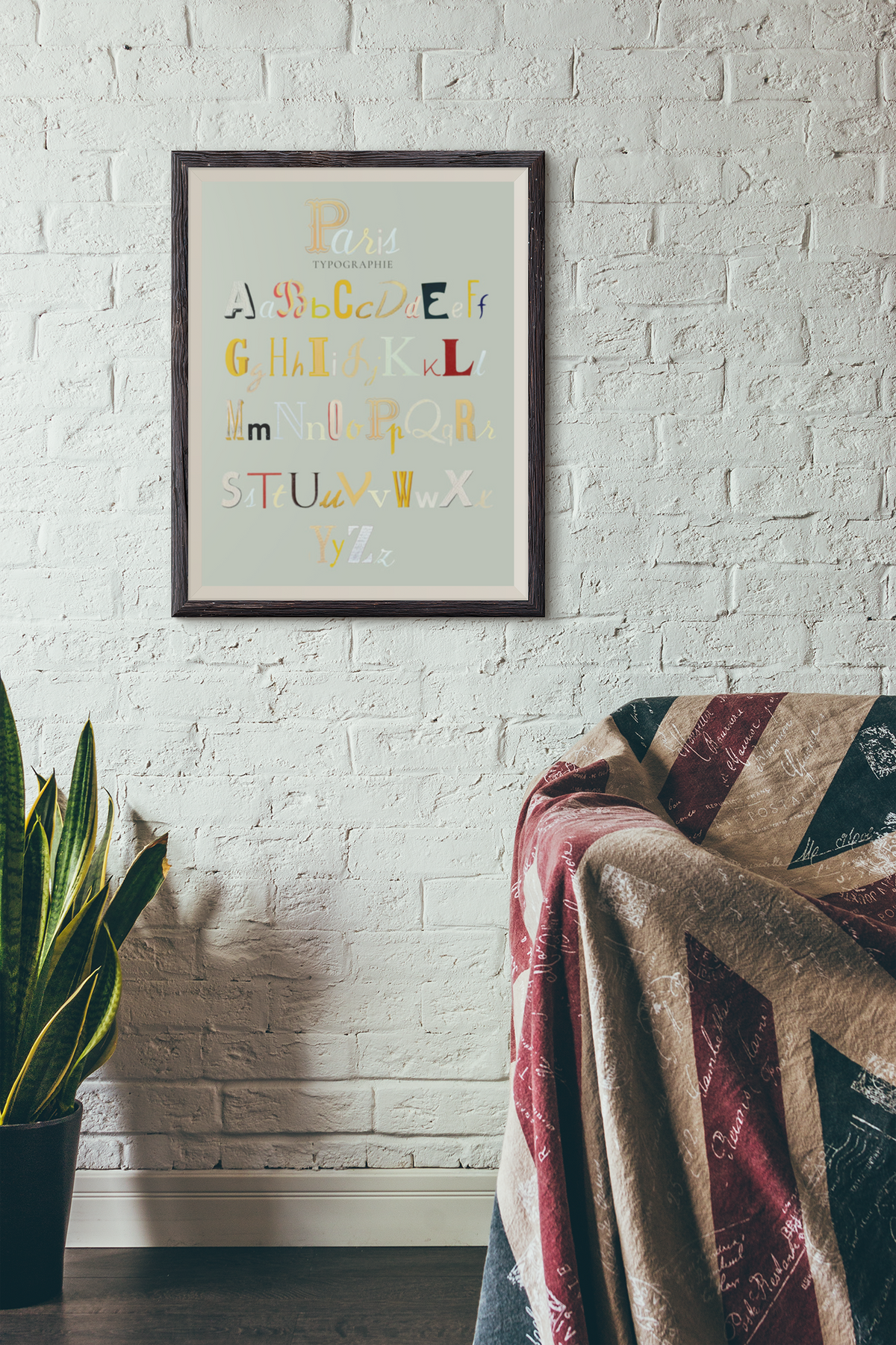 Framed alphabet wall art hanging on a white brick wall above a sofa with a Union Jack throw blanket, with a potted plant at left.